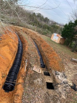Drain field install on a sloped lot: two trenches in orange clay with black chamber lines and open septic tank lids